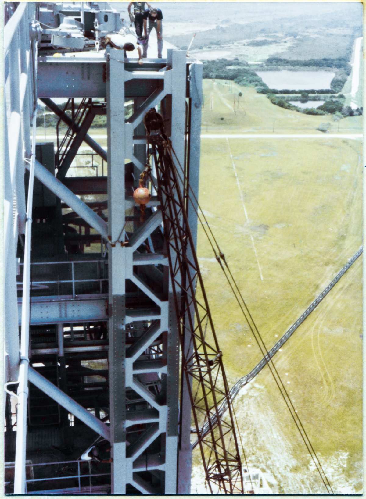 Image 080. Wade Ivey (dark blue shirt, holding blue hardhat in his right hand), owner of Ivey's Steel Erectors, stands on the checkerplate which defines the top of the Fixed Service Structure at elevation 300'-0”, in this image taken from the far end of the Hammerhead Crane Boom at Space Shuttle Launch Complex 39-B, Kennedy Space Center, Florida, and leans out past the edge of the checkerplate in order to see precisely where his team of Union Ironworkers from Local 808 have positioned the GOX Arm Hinges Support Strongback. The Strongback is not quite at its final location, where it will be welded and bolted to the Perimeter Framing of the FSS, and great care must be taken using a mobile crane with just under 250 feet of boom, maneuvering horizontally and vertically to sub-eighth-inch tolerance, in conjunction with rigging attached directly to the FSS itself, to get the Strongback EXACTLY where it belongs, where it will be fastened immovably in place, thus irrevocably locating where the GOX Arm itself will be positioned for its servicing work at the Gaseous Oxygen Vent on top of the Space Shuttle's External Tank. This is exceedingly close-tolerance work, and it takes the best in the business to execute it, and Wade Ivey along with his whole workforce of Union Ironworkers were the best in the business. Photo by James MacLaren.